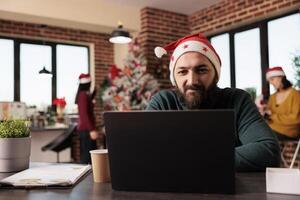 Smiling company employee checking startup project report on laptop while working in office at christmas eve. Caucasian man in santa hat using computer at festive decorated workplace photo