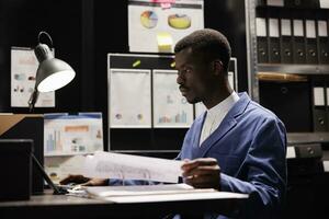 Businessman analyzing administrative files, reviewing management report in storage room. Bookkeeper in formal suit working late at night at accountancy record in corporate depository photo