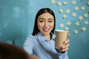 Smiling woman showing disposable coffee cup while looking towards cell phone camera. Female employee enjoying first coffee of the day to give her energy for a long day at work. photo