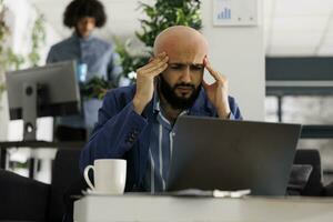 Start up product manager with headache working to meet deadlines in business office. Arab company entrepreneur suffering from migraine and rubbing temples at workplace in coworking space photo