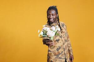 Portrait of african american man holding white flowers bouquet, presenting surprise for girlfriend at camera in studio over yellow background. Romantic boyfriend posing for valentine's day photo