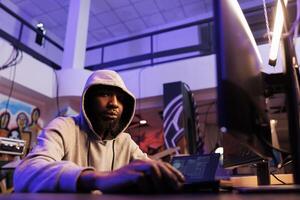 Hacker coding internet virus malicious software on computer portrait. African american man in hood doing ddos attack illegal activity and breaking law while looking at camera photo