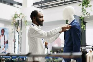 Clothes retail shop african american assistant dressing mannequing in formal suit to showcase apparel in stock. Shopping mall fashion department man employee putting male blue jacket on model photo