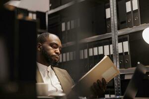 Exhausted african american detective solving complex crime case and reading folder with police reports. Tierd investigator working late, studying archival records information photo