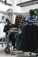 Clothing store smiling diverse employee and client in wheelchair chatting while choosing casual shirt together. Shopping mall cheerful asian seller and arab customer with disability exploring garment photo