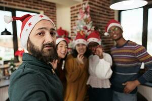 Diverse workers taking smartphone selfie at christmas corporate party in decorated office. Colleagues in santa hats posing for mobile phone photo while celebrating new year at workplace