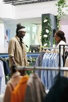 African american man and woman couple shopping in clothing store. Buyers choosing trendy outfit in fashion boutique while standing and talking near shirts hanging on racks photo