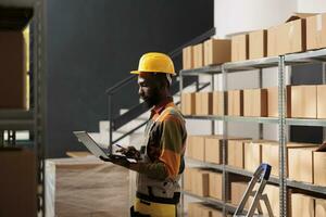 African american worker holding laptop computer, analyzing products checklist in warehouse. Storage room employee wearing protective overall while preparing customers orders before delivery goods photo