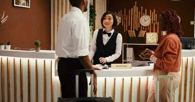 Friendly asian receptionist assisting guests with booking reservation, inviting them to wait in stylish cozy hotel lounge. Happy guests helped by bellboy with their luggage photo