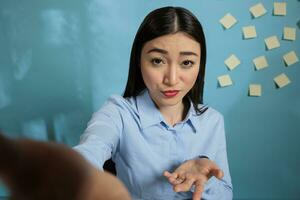 Asian woman in video call with mobile phone looking at the screen while asking for an explanation of what is happening by gesturing with hand. Employee in remote communication talking to coworkers. photo