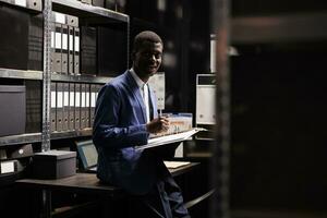 Private detective working at confidential case, analyzing files with evidence about crime scene in arhive room. African american police officer checking criminology report late at night photo