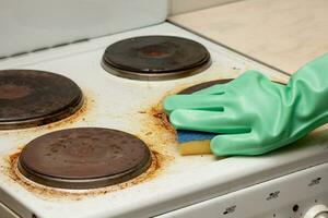Woman hand in glove remaining burnt stains on dirty electric stove scrubbing using sponge photo