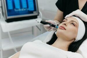 Shot of a beautiful young woman on a facial dermapen treatment at the beauty salon. Dermatologist in medical gloves doing hydro peeling procedure. Close up facial treatment. photo