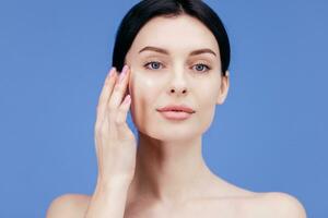 Close-up beauty portrait of young woman with smooth healthy skin, she gently touches her face with her fingers on light grey background and smiles. Skincare concept photo