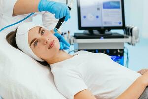 Cosmetology clinic. Professional female cosmetologist doing hydrafacial procedure while being a work. Attractive nice woman lying on the medical bed while having beauty procedures photo