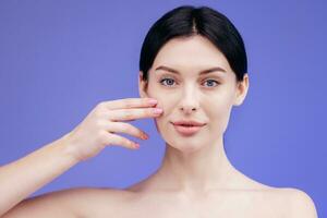 Close-up beauty portrait of young woman with smooth healthy skin, she gently touches her face with her fingers on light grey background and smiles. Skincare concept photo