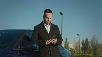 An elegant businessman in a coat stands next to his premium car and uses a smartphone to solve household and work issues. video