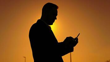 silhueta do uma homem contra a pôr do sol céu. uma homem usa uma Smartphone, tipos mensagens dentro formulários e social redes video