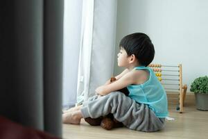 triste asiático chico se sienta con su cabeza abajo y mira fuera el puerta a su favorito muñeca. foto