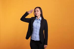 Patriot filipino woman doing military salute with hand over forehead, promotiing honor and respect in front of camera over yellow background Proud adult showing army soldier hello gesture photo