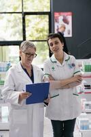 dos sonriente mujer farmacia especialistas en farmacia retrato. mayor farmacéutico empleado participación prescripción portapapeles y joven boticario trabajador mirando a cámara foto