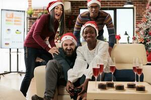 Excited employees holding gamepads and enjoying video game in festive decorated office on xmas eve. Cheerful diverse company coworkers fighting in videogame at new year holiday party photo