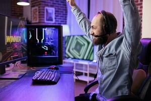 Gamer winning intense singleplayer spaceship arcade racing videogame on capable gaming computer system at home. Man throwing hands in air from happiness after finally passing difficult game level photo