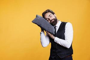 Sleepy butler sleeping on pillow, taking a nap while he in standing in studio. Young exhausted overworked person falling asleep against yellow background, working in restaurant industry. photo