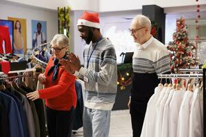 Employee showing items to senior couple shopping for family gifts, looking for presents in festive clothing store. African american retail assistant helping elderly people to choose perfect clothing. photo