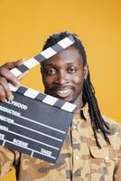 Portrait of producer holding clapperboard to cut scenes in movie making industry. African american adult presenting chalkboard to camera and smiling while having filmmaker object. Cinema concept photo