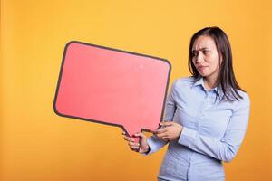 Smiling filipino woman pointing at empty red speech bubble with copy space, showing advertising mock up. Cheerful young adult holding dialog frame, person with communication cloud in studio photo