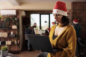 Woman project manager wearing santa hat managing tasks on laptop while standing in winter festive office. Employee holding portable computer and working for deadline at christmas eve photo