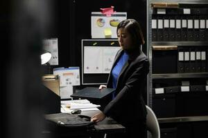 Asian woman private investigator unpacking laptop before beginning criminal investigation case. Detective in archive room office filled with criminology folders on bookshelves photo