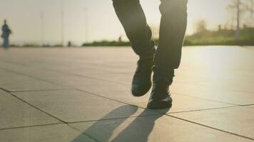 een onherkenbaar Mens in klassiek schoenen en jeans wandelingen aan de overkant de vierkant. detailopname van wandelen voeten tegen de backdrop van de instelling zon. video