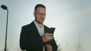 elegante urbano uomo nel un' cappotto e bicchieri nel il parco. un' uomo d'affari usi un' smartphone per opera nel applicazioni e sociale reti. video