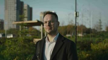 un satisfecho elegante hombre en lentes camina mediante un ciudad parque. el hombre es vistiendo un elegante Saco y camisa. video