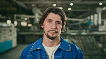 Portrait of a young worker in a spacious workshop at a manufacturing plant. A friendly man in a blue uniform looks at the camera. video