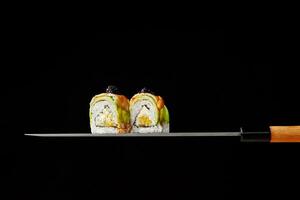 Two uramaki rolls with cream cheese, mango, avocado and shrimps on blade of Japanese knife on black background photo