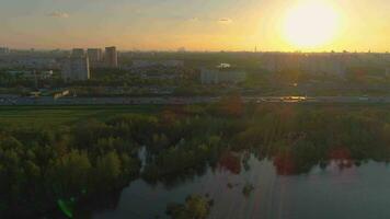 Flying over the lake, trees, cars traffic on highway and city buildings at colorful sunset against the sun. Aerial view. video