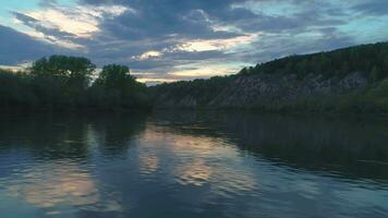 veloce volante per il giusto al di sopra di il fiume a tramonto. rocce con verde alberi su un' lungo il fiume. colorato cielo. video