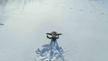 Young cheerful woman is falling in snow and is making snow angels. Slow motion. Aerial view, flying forward video