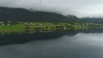 pequeño pueblo y reflexión en lago. verde paisaje de Noruega en nublado verano día. aéreo vista. zumbido es volador oblicuo y ascendente video