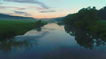 Flying forward over river in mist at sunrise. Green trees, meadow and sky reflect in water. Aerial view. video