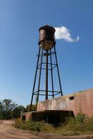 A beautiful water tower is set around an abandoned area. This rusty metal structure stands tall against a blue sky. photo