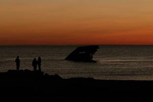 Sun setting on the Delaware Bay. The pretty red colors like the sky is on fire. The sunken ship of Cape May sitting in the ocean. People standing on the large black rocks of the jetty of the beach. photo