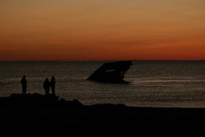 Sun setting on the Delaware Bay. The pretty red colors like the sky is on fire. The sunken ship of Cape May sitting in the ocean. People standing on the large black rocks of the jetty of the beach. photo