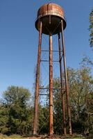 un hermosa agua torre es conjunto alrededor un abandonado área. esta oxidado metal estructura soportes alto en contra un azul cielo. foto