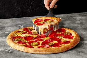 Female hand holding serving paddle with slice of pepperoni pizza with melted cheese and jalapeno photo