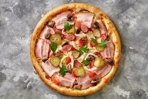 Country style pizza with chicken, bacon, hunting sausages, pickled cucumbers, mushrooms, vegetables and cheese on gray surface photo