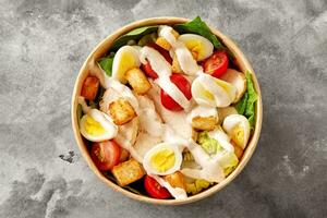 Paper bowl with Caesar salad of romaine lettuce, cherry tomatoes, quail eggs, croutons, chicken and sauce on gray surface photo